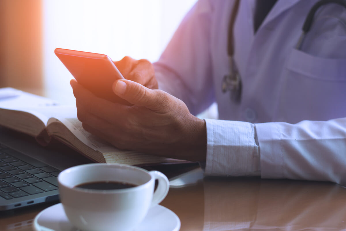Male doctor hand holding and using mobile smart phone work on laptop computer text book and cup of coffee on the wooden desk in medical room at clinic or hospital | コーヒーステーション