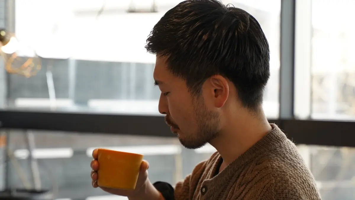 茶色いセーターを着た男性が、オレンジ色のカップを手に持ち、目を閉じて香りを確かめているかのように見える。窓からの光が差し込む穏やかな空間でリラックスしている様子。