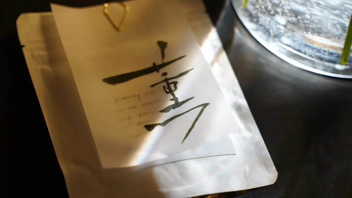 白いパッケージに墨で「重」と書かれた和紙のラベルが金色のクリップで留められています。背景には水泡が輝くグラスと緑の茎が見え、光が差し込む静かで上品な雰囲気の画像です。