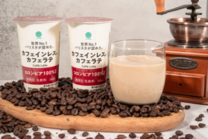 Two cafe latte cartons with Japanese text sit on a bed of roasted coffee beans beside a glass of latte and a vintage hand cranked grinder in the background