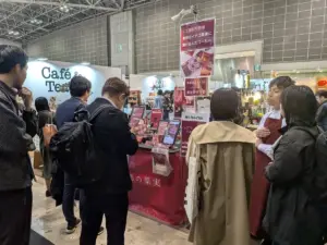 Crowd of visitors at a pink themed exhibition booth a vendor in a red apron explaining products to attendees while display shelves line the table