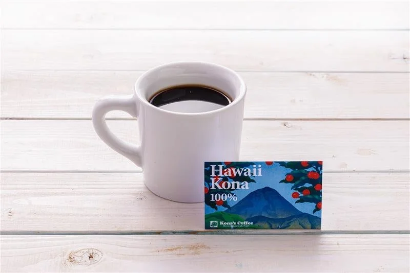 白い木目のテーブルに、白いマグカップに入ったホットコーヒーと、ハワイコナコーヒーのパッケージが置かれています。