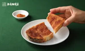 0106f3ef6432484ba94ee9986d9762c9 | コーヒーステーション Toasted bread slices on a white plate with a hand lifting one piece a small bowl of red spice on a green table nearby