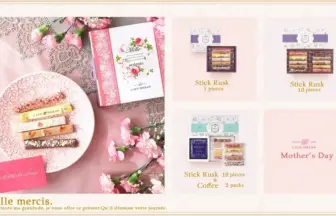 Pink-themed Mother’s Day gift layout with a Mille mercis box, lace tablecloth, and pink flowers surrounding ceramic plate with assorted stick rusk pastries.