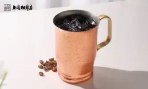 07ad6d78e90eeb06c6abb4a56969d38d | コーヒーステーション Copper mug with a black interior filled with ice condensation on the outside and a gold handle on a white surface with coffee beans nearby