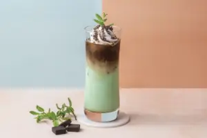 Tall iced matcha latte with coffee crema topped with whipped cream and chocolate crumbs mint leaf on a marble coaster with chocolate pieces nearby