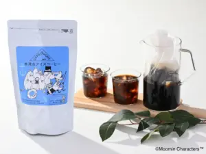 White coffee bag with a blue Moomin Characters label sits beside two glasses of iced coffee and a glass carafe on a wooden board with green leaves nearby