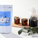 White coffee bag with a blue Moomin label beside two glasses of iced coffee and a glass pitcher on a wooden board product shot for iced coffee mix