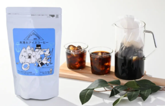 White coffee bag with a blue Moomin label beside two glasses of iced coffee and a glass pitcher on a wooden board product shot for iced coffee mix