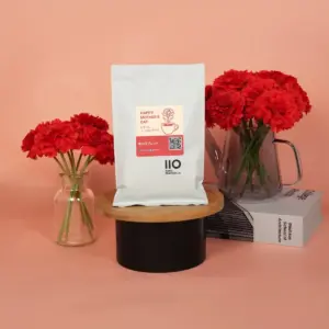 18e708280119187c928a28c734f2889f | コーヒーステーション White coffee bag on a round wooden stand with red carnations in glass vases against a pink backdrop a book nearby showing a Mothers Day label in pinkred
