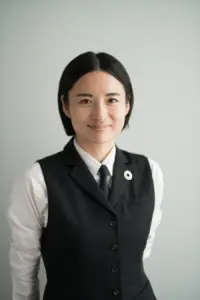 19c311a65dd68559875dfd825ce51ffb | コーヒーステーション Portrait of a young Asian woman in a black vest white shirt and navy tie smiling at the camera with a circular lapel pin visible on her left chest