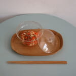 Clear glass bowl with carrot and tofu salad on a wooden tray lid beside it set on a pale blue table with chopsticks below