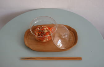 Clear glass bowl with carrot and tofu salad on a wooden tray lid beside it set on a pale blue table with chopsticks below
