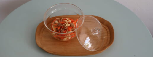 Clear glass bowl with carrot and tofu salad on a wooden tray lid beside it set on a pale blue table with chopsticks below