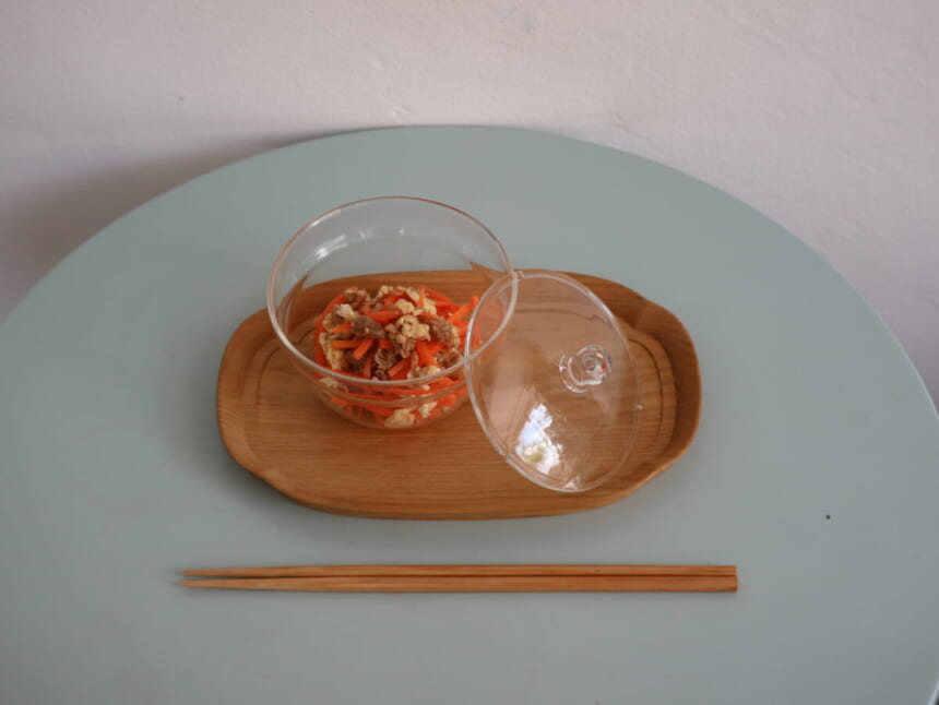 Clear glass bowl with carrot and tofu salad on a wooden tray lid beside it set on a pale blue table with chopsticks below