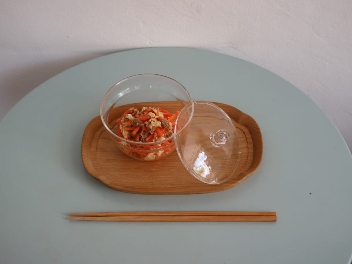 Clear glass bowl with carrot and tofu salad on a wooden tray lid beside it set on a pale blue table with chopsticks below