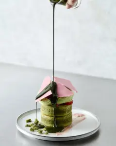 Stack of green matcha macarons topped with pink chocolate shards on a white plate with dark green sauce streaming over them