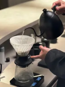 1c6da25fb3b660c8a542d43b25d9963f | コーヒーステーション Barista pours hot water from a black gooseneck kettle into a paper filter in a glass coffee dripper atop a digital scale