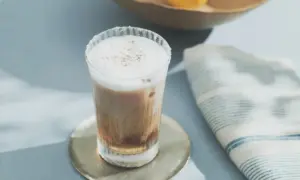 Layered coffee drink in a clear glass with foam on top on a round metal coaster beside a blue striped napkin