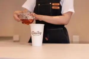 Barista pours tea from a glass teapot into a white disposable cup labeled Daily Tea  Coffee