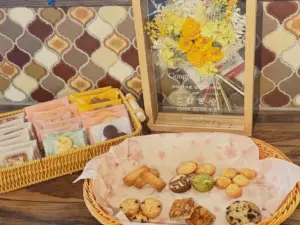 Basket of assorted cookies on a wooden table with wrapped treats nearby and a framed yellow flower arrangement in the background