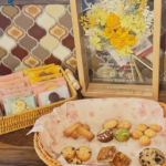 Basket of assorted cookies and pastries on a wooden table with a framed yellow flower bouquet in the background