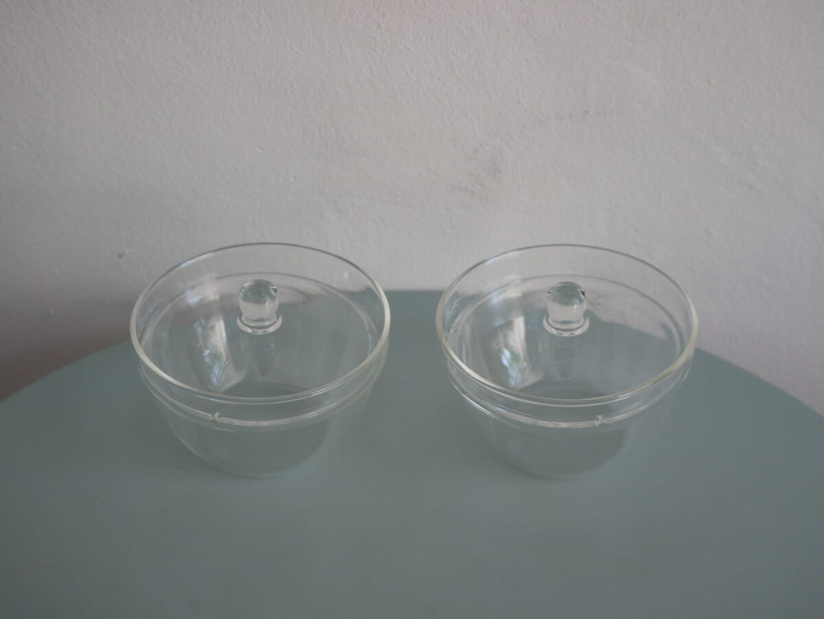 Two clear glass bowls with lids on a light blue table surface