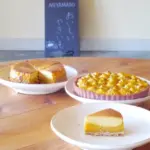 Three desserts on a wooden table: a cheesecake slice on a white plate, a round nut-topped tart, and a slice of layered cake, with a chalkboard sign in the background.
