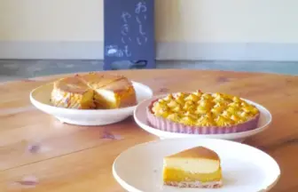 Three desserts on a wooden table: a cheesecake slice on a white plate, a round nut-topped tart, and a slice of layered cake, with a chalkboard sign in the background.