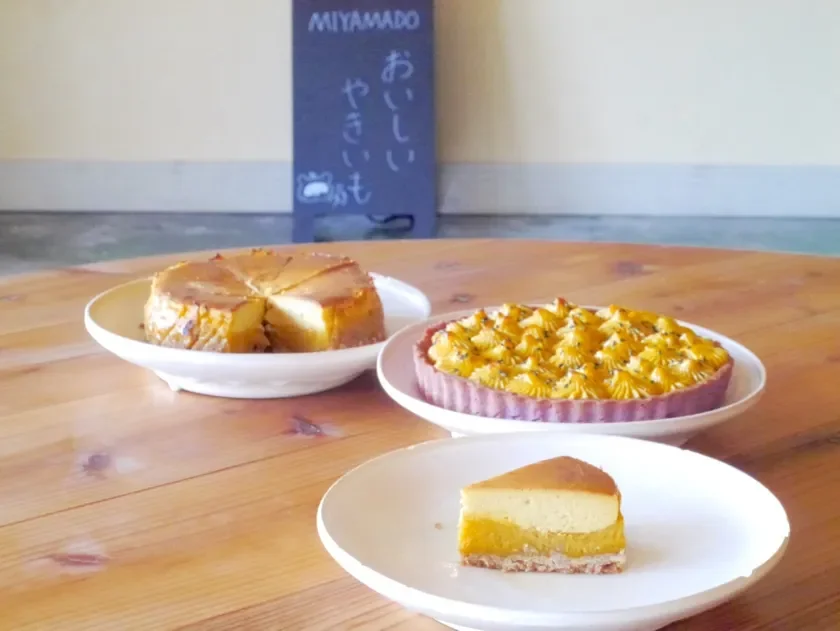 Three desserts on a wooden table a cheesecake slice on a white plate a round nut topped tart and a slice of layered cake with a chalkboard sign in the background