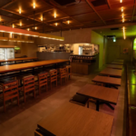 Warm modern restaurant interior with long wooden tables and stools open kitchen in back and booths along the right wall under amber lighting