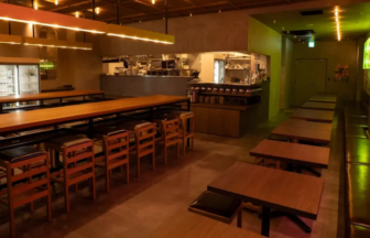 Warm modern restaurant interior with long wooden tables and stools open kitchen in back and booths along the right wall under amber lighting