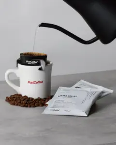 2ce5974c17303d23bf62e2e8f7cb83dc | コーヒーステーション White mug labeled PostCoffee with a drip bag and grounds as hot water flows from a black kettle into it