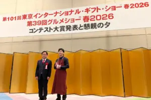 Two presenters on a stage with gold folding screens one in a suit with a red rosette the other in a maroon apron holding a trophy beneath a large Japanese banner
