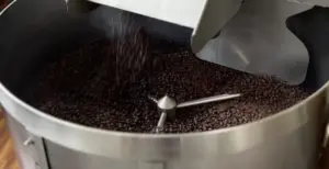 Dark roasted coffee beans tumbling in a stainless steel commercial roaster with a mixing arm overhead