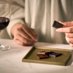 Persons hand holds a small dark chocolate square over a tray of assorted chocolates on a table with a glass of red wine nearby