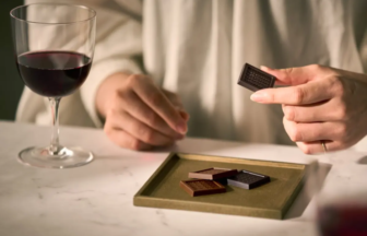 Persons hand holds a small dark chocolate square over a tray of assorted chocolates on a table with a glass of red wine nearby