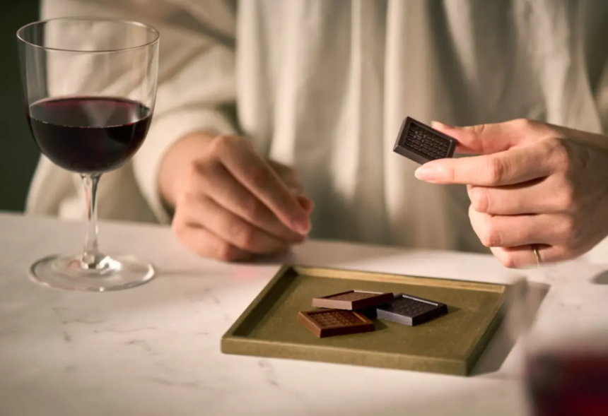 Persons hand holds a small dark chocolate square over a tray of assorted chocolates on a table with a glass of red wine nearby