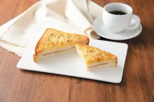 36a0277b833c61f9ef8e8396b9ee1ad1 | コーヒーステーション Two slices of cheesy layered ham and cheese sandwich on a white plate with a cup of coffee nearby on a wooden table