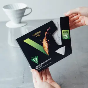 Close up of a hand holding a Hario V60 coffee filter box on a gray surface with a white ceramic dripper visible in the background