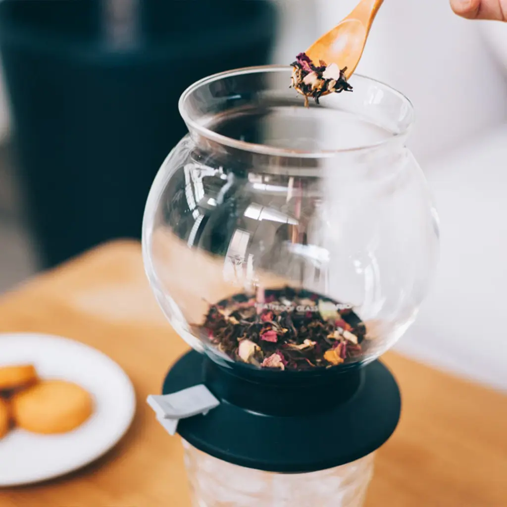 Glass teapot with loose tea leaves on a black base a wooden spoon hovering above to add tea cookies on a plate visible in the background
