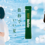 Woman standing by a sunny sea holding a small glass of coffee with a vertical green Blendy sachet in the center and a hand holding a glass on the right Japanese slogan text nearby