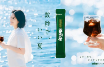 Woman standing by a sunny sea holding a small glass of coffee with a vertical green Blendy sachet in the center and a hand holding a glass on the right Japanese slogan text nearby