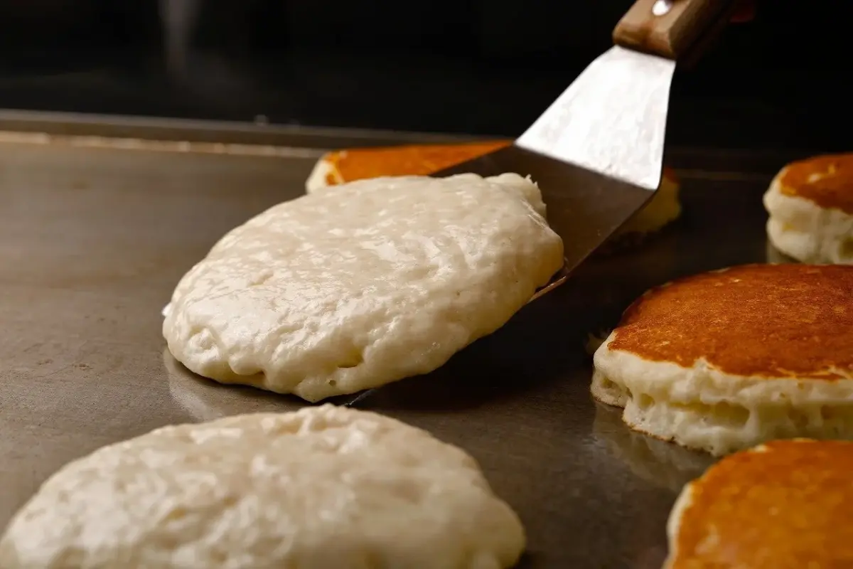 熱い鉄板の上でパンケーキが焼かれている様子です。フライ返しでひっくり返されようとしている白い生地と、すでに焼き色のついたパンケーキが複数見えます。