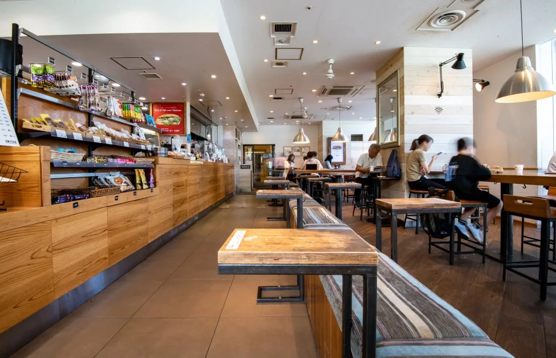 カフェの店内の様子。木目調のカウンターには軽食やパンが陳列されており、奥にはイートインスペースが広がっています。数人の客がそれぞれの席で食事や休憩を楽しんでおり、リラックスした雰囲気が漂っています。
