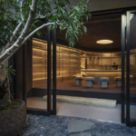 Open glass doors reveal a sleek dimly lit bar with a curved counter and beige stools inside a modern interior