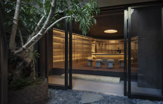 Open glass doors reveal a sleek dimly lit bar with a curved counter and beige stools inside a modern interior