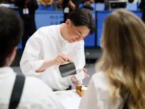Man in a white shirt tamping espresso into a black cup during a coffee competition with spectators in the background in a cafe or event setting