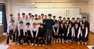Group photo in a classroom showing a man in the center surrounded by students in white shirts and navy uniforms posing for the picture