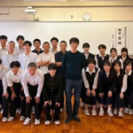 Group of students in uniforms posing with a man in the center in a classroom setting smiling for a class photo